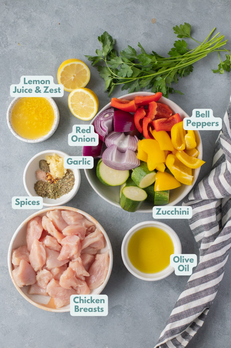 Grilled Greek Chicken Kabobs Ingredients in small white bowls on a grey background: boneless skinless chicken breasts cut into 1 inch, fresh vegetables (bell peppers, red onion, zucchini), extra virgin olive oil, lemon juice, lemon zest, minced garlic, dried basil, dried oregano, dried thyme, salt and pepper, red pepper flakes