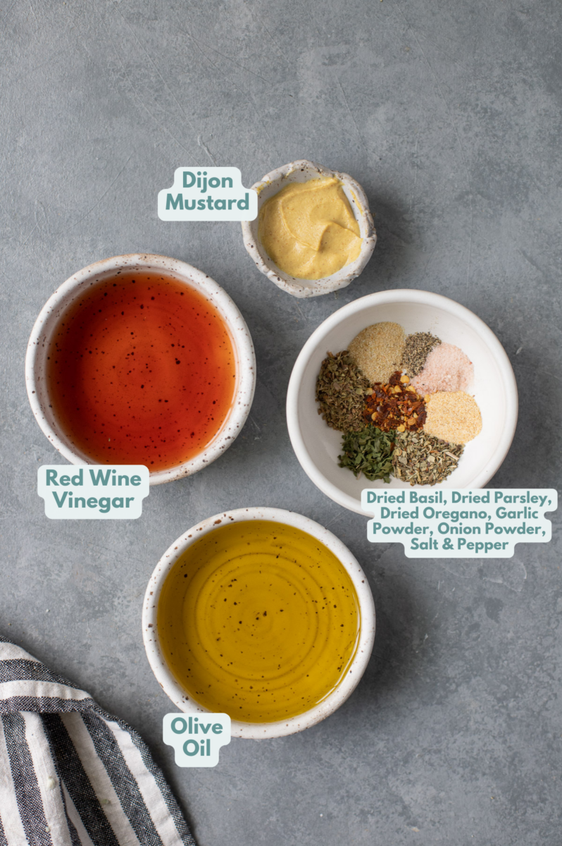 Ingredients for Homemade Italian Dressing in small white bowls on a grey background: extra virgin olive oil, red wine vinegar, dijon mustard, dried basil, dried parsley, dried oregano, garlic powder, onion powder, salt, pepper, red pepper flakes