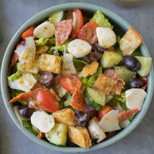An overhead shot of a chopped italian salad in a bowl with a clear jar of italian dressing on the side on a blue background