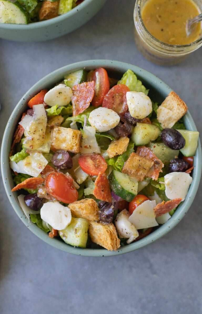 An overhead shot of a chopped italian salad in a bowl with a clear jar of italian dressing on the side on a blue background