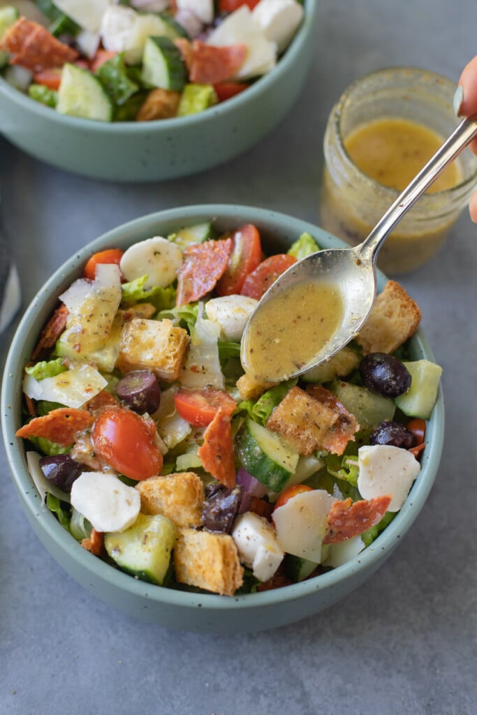 A chopped italian salad in a bowl with a spoon drizzling more dressing on top with a clear jar of italian dressing on the side on a blue background