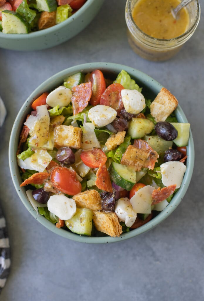 An overhead shot of a chopped italian salad in a bowl with a clear jar of italian dressing on the side on a blue background