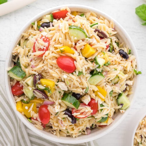 An overhead shot of a white bowl of orzo salad on a white background with tomatoes, basil and a towel on the side