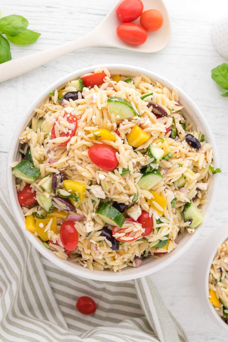 An overhead shot of a white bowl of orzo salad on a white background with tomatoes, basil and a towel on the side