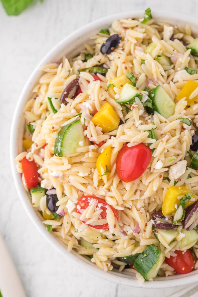 An overhead shot of a white bowl of orzo salad on a white background with tomatoes, basil and a towel on the side