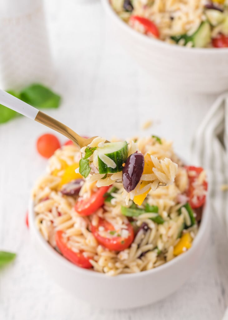 A fork with a bite of orzo salad with a small white bowl of orzo salad in the background