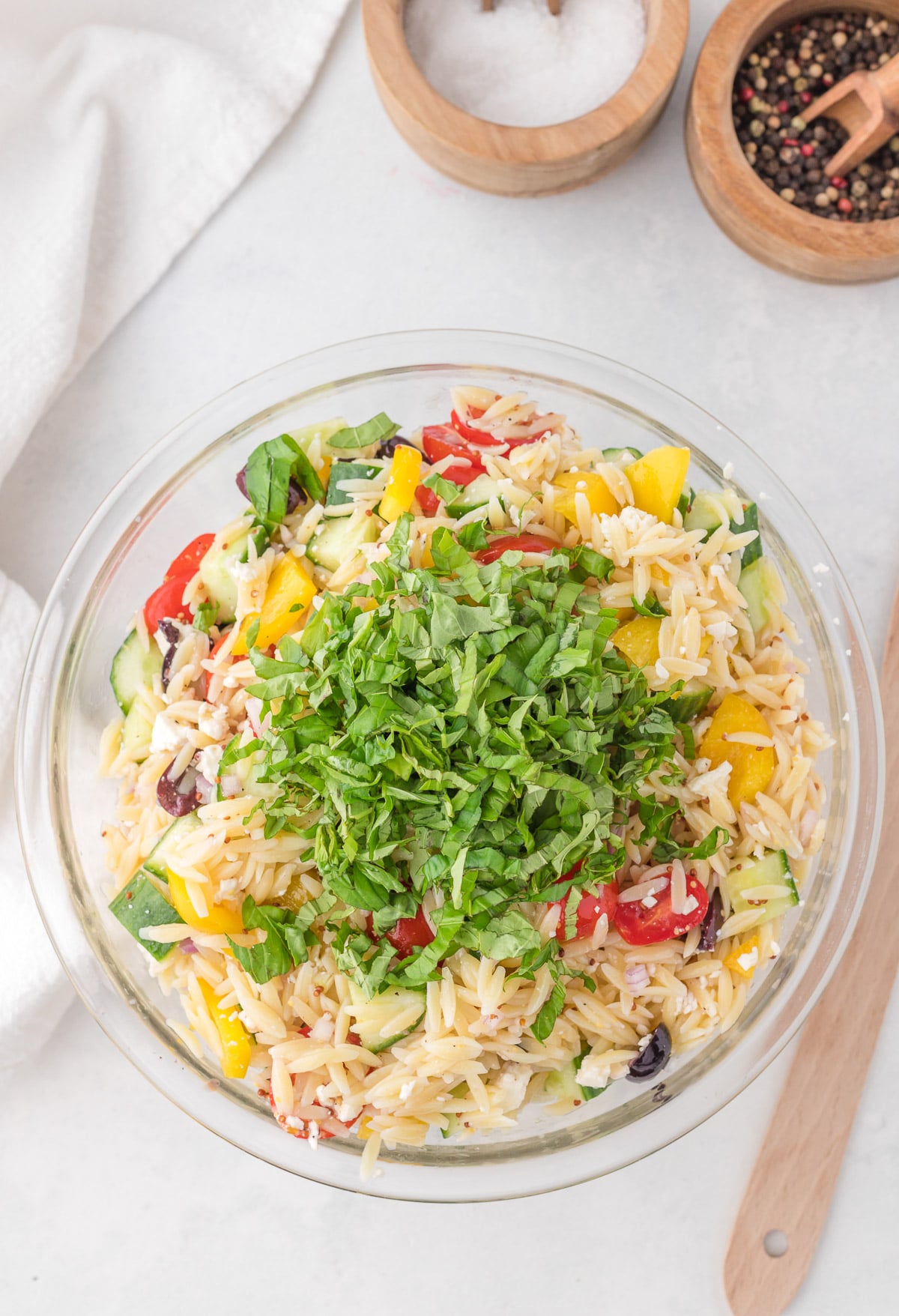 Orzo salad in a clear bowl with fresh chopped basil on top before being mixed in 