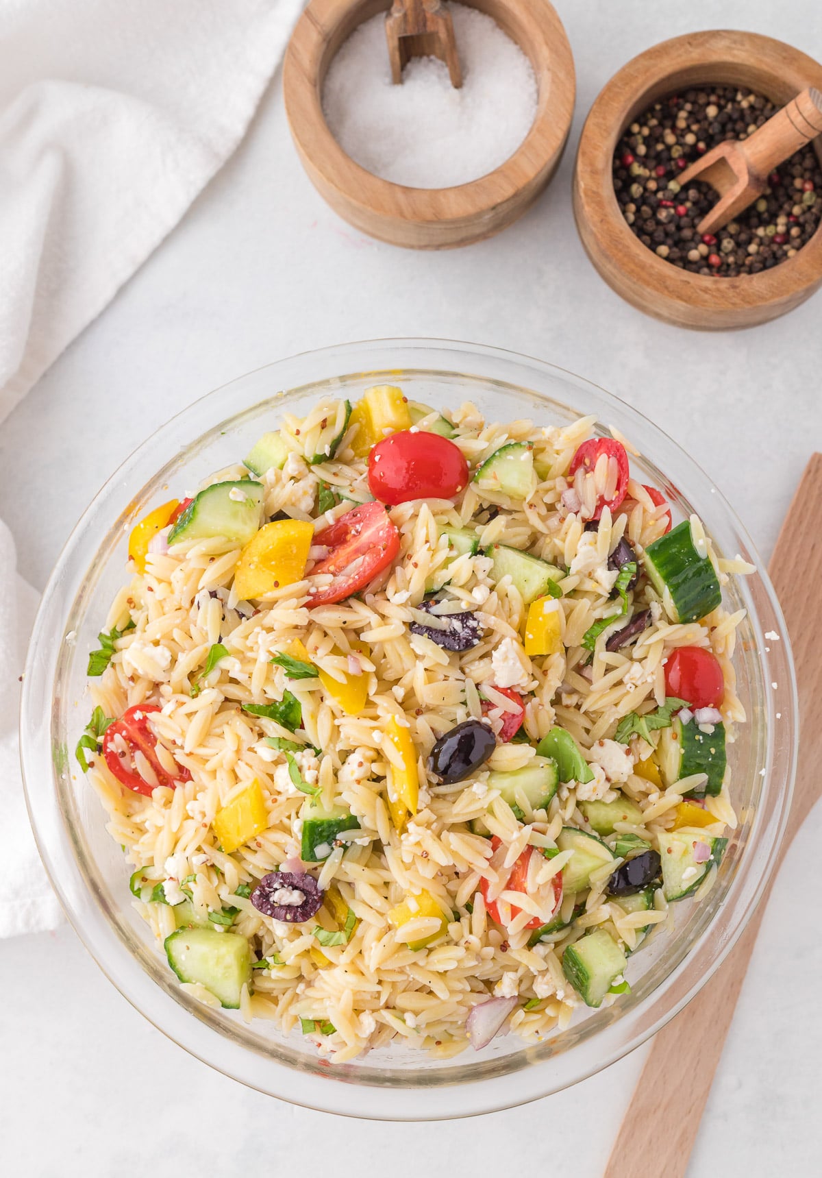An overhead shot of a white bowl of orzo salad on a white background with salt and pepper on the side