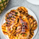 an overhead shot of a white plate of pineapple grilled chicken with a pineapple on the side on a grey background