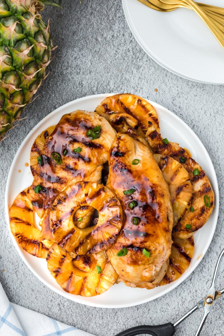 an overhead shot of a white plate of pineapple grilled chicken with a pineapple on the side on a grey background