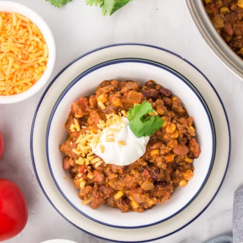 An overhead shot of a white bowl with pumpkin chili with cilantro, cheese, and limes on the side
