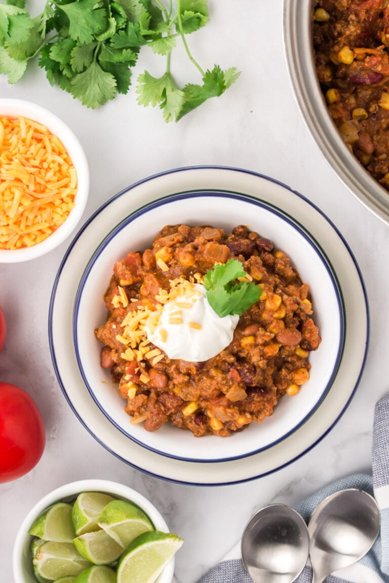 An overhead shot of a white bowl with pumpkin chili with cilantro, cheese, and limes on the side
