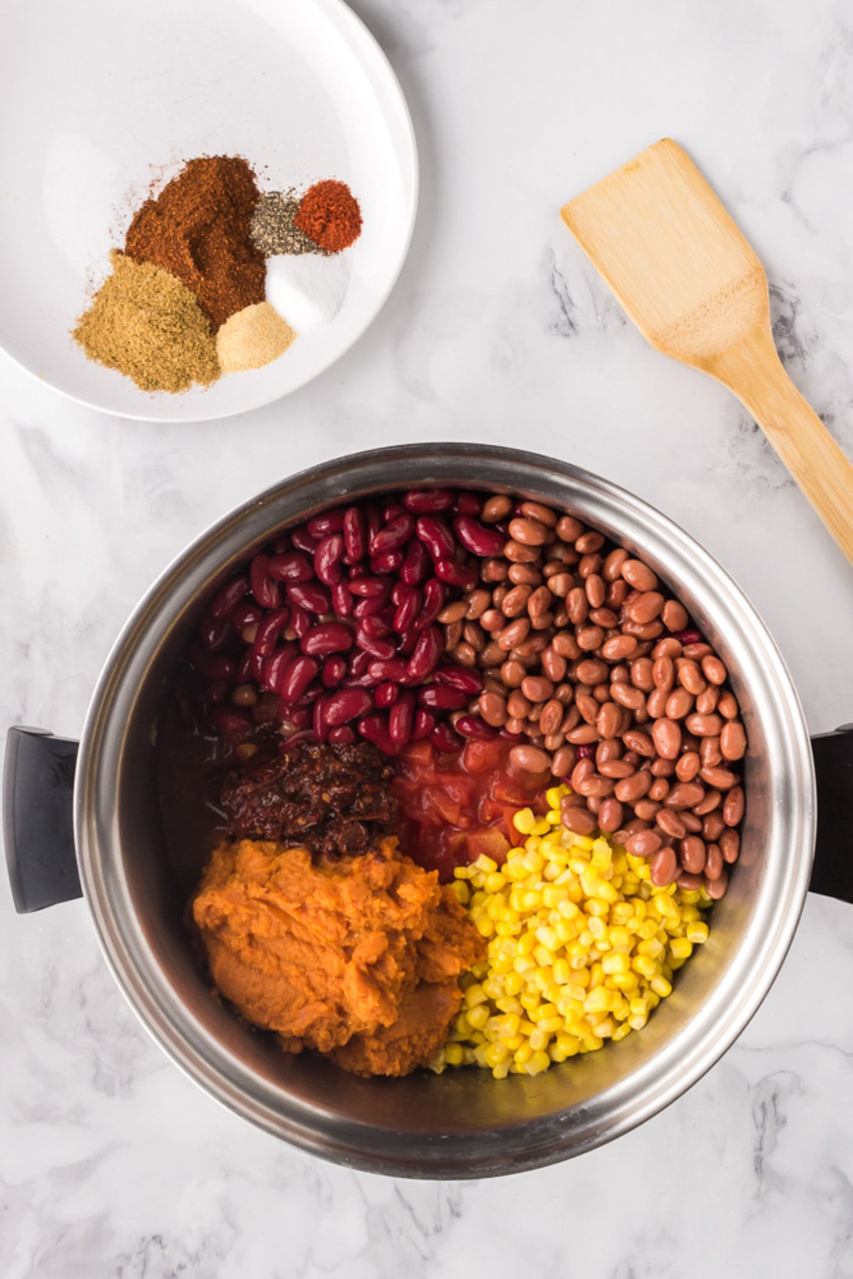 All ingredients for pumpkin chili in a pot before stirring with a wooden spoon and spices on the side