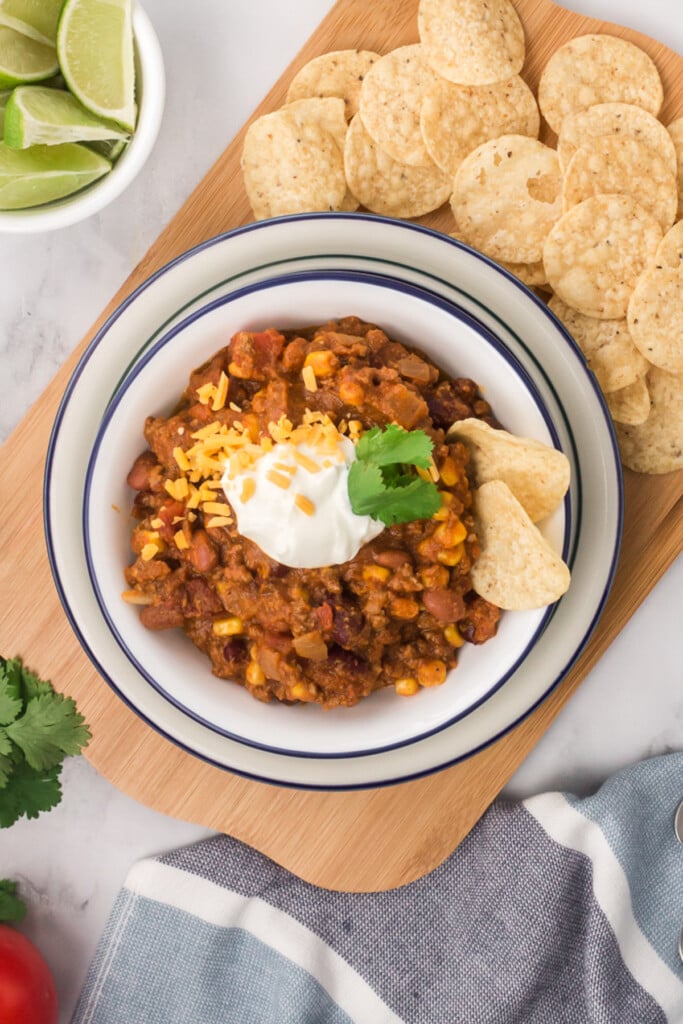 An overhead shot of a white bowl with pumpkin chili with limes and tortilla chips on the side