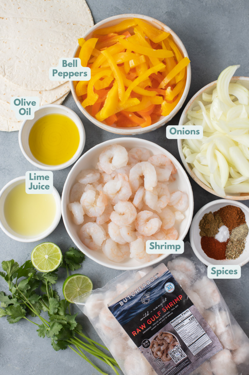 An overhead image of shrimp fajitas ingredients in small white bowls on a grey background: shrimp, olive oil, lime juice, paprika, ground cumin, salt, chili powder, oregano, pepper, bell peppers, onions, flour tortillas 