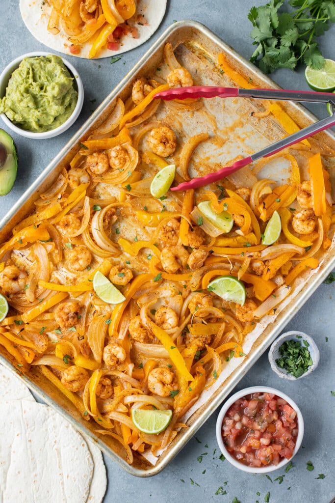 Sheet pan shrimp fajitas on a baking sheet with lime wedges and with tacos, salsa, and guacamole on the side
