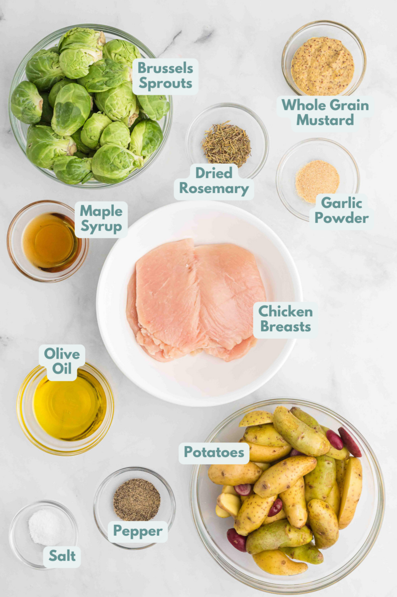 Ingredients for maple mustard chicken laid out on a grey background in small bowls: chicken, potatoes, brussels sprouts, maple syrup, whole grain mustard, salt, pepper, garlic powder, rosemary, olive oil
