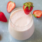 An overhead shot of a strawberry smoothie in a clear glass with a strawberry on the rim with strawberries in the background