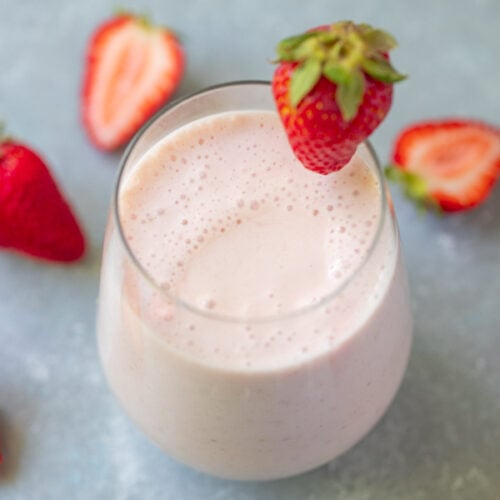 An overhead shot of a strawberry smoothie in a clear glass with a strawberry on the rim with strawberries in the background