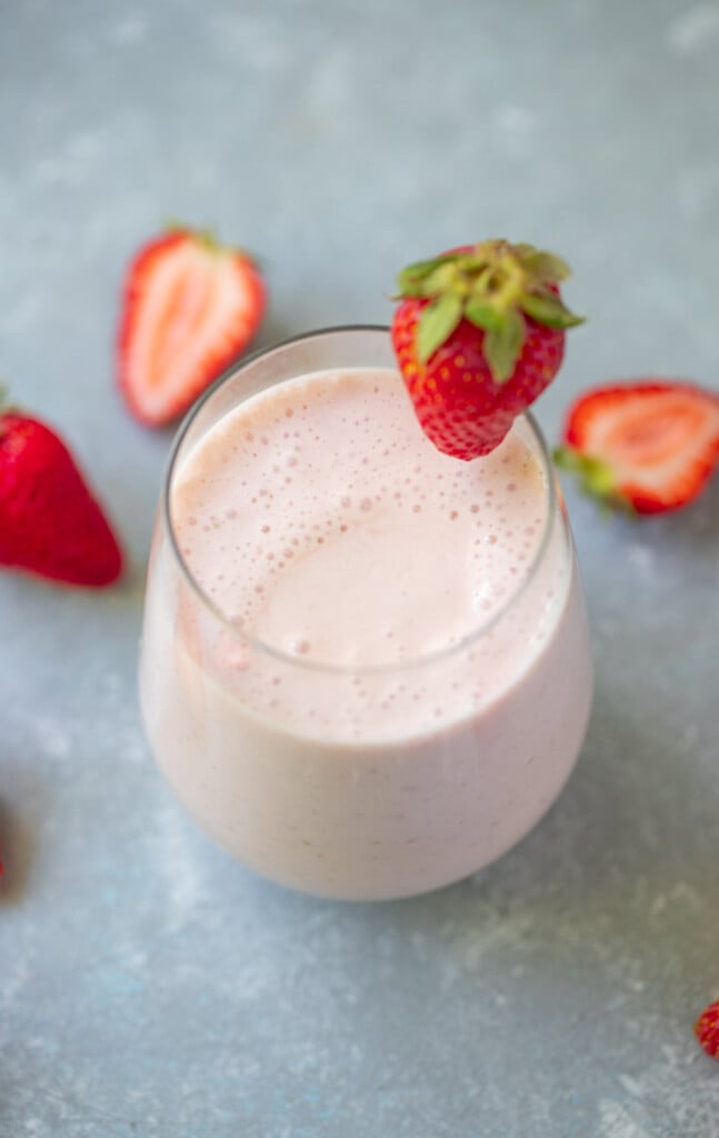 An overhead shot of a strawberry smoothie in a clear glass with a strawberry on the rim with strawberries in the background