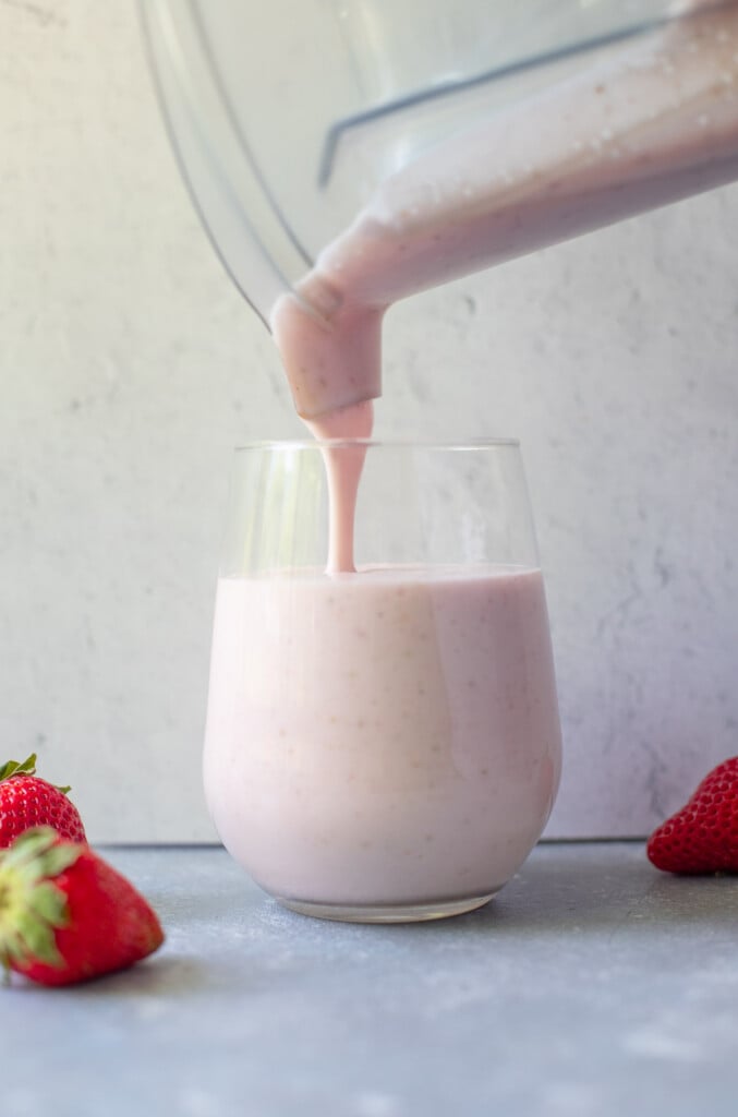 Strawberry smoothie being poured into a clear glass from a blender with strawberries on the side