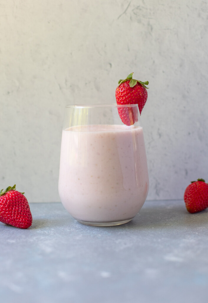 A smoothie with strawberries in a clear glass with a strawberry on the rim with strawberries on the side