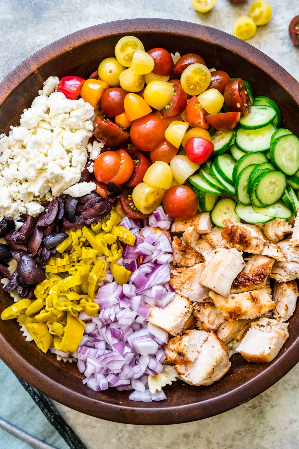 a large wooden bowl with ingredients for greek chicken pasta salad before dressing is poured on and before mixing