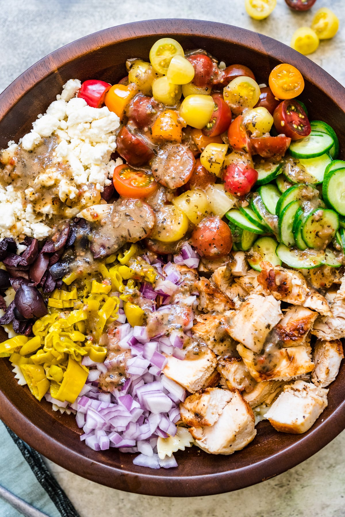 a large wooden bowl with ingredients for greek chicken pasta salad with dressing poured on top but before mixing