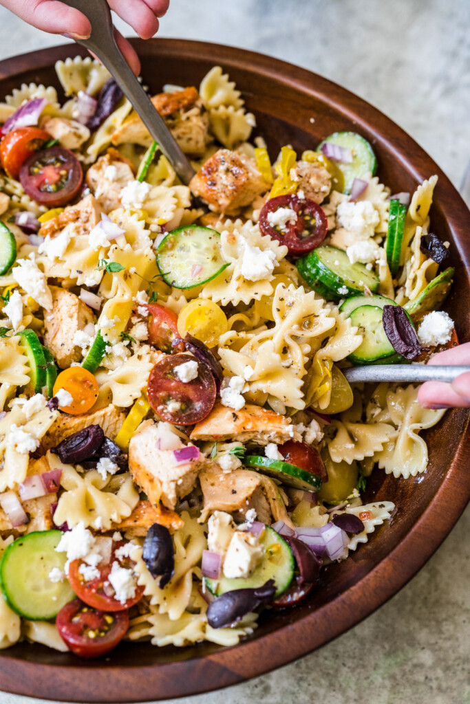 A wooden bowl of greek chicken pasta salad being tossed with two utensils