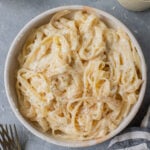 A bowl of healthy alfredo on a grey background with forks, cheese, sauce and a linen across it.