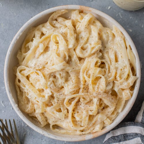 A bowl of healthy alfredo on a grey background with forks, cheese, sauce and a linen across it.