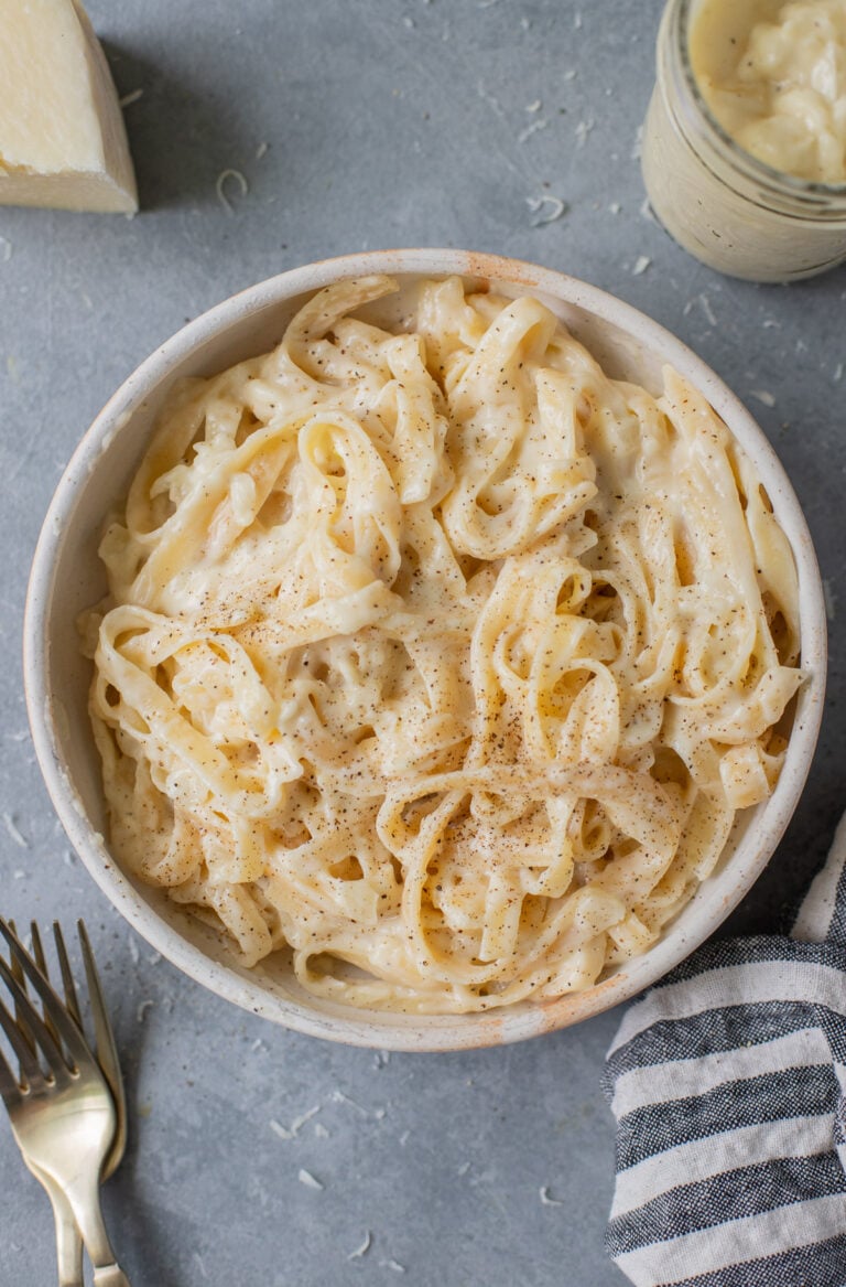 A bowl of healthy alfredo on a grey background with forks, cheese, sauce and a linen across it.