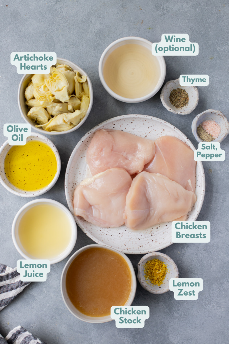 Ingredients for Artichoke Chicken on a grey background in small white bowls: olive oil, chicken breasts, salt, pepper, canned artichoke hearts, lemon juice/zest, chicken broth, thyme and white wine