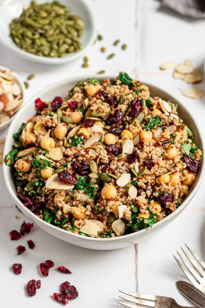 A white bowl of quinoa kale salad with cranberries and pepitas on the side on a white background