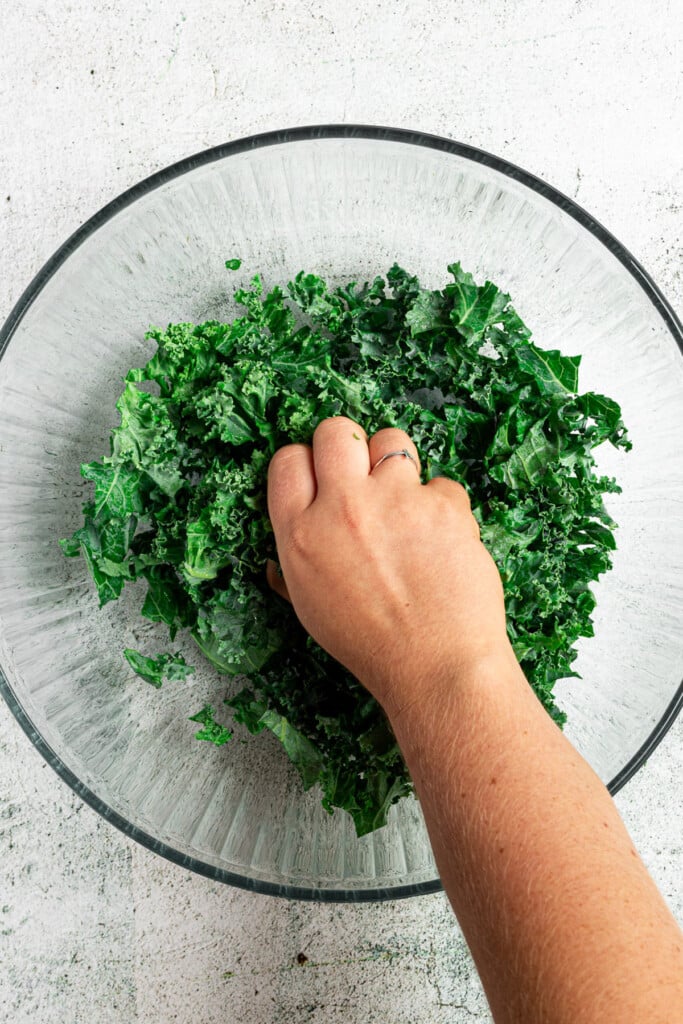 A clear bowl filled with kale with a hand massaging the kale for a kale quinoa salad