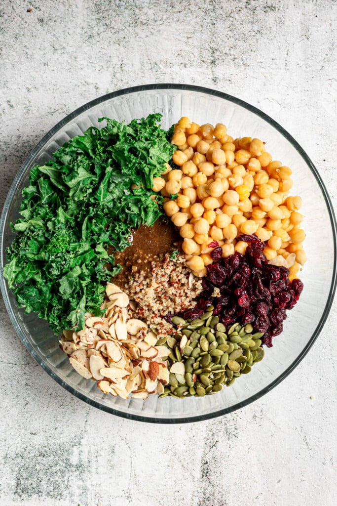 All ingredients for kale quinoa salad in a large clear bowl before being mixed on a grey background