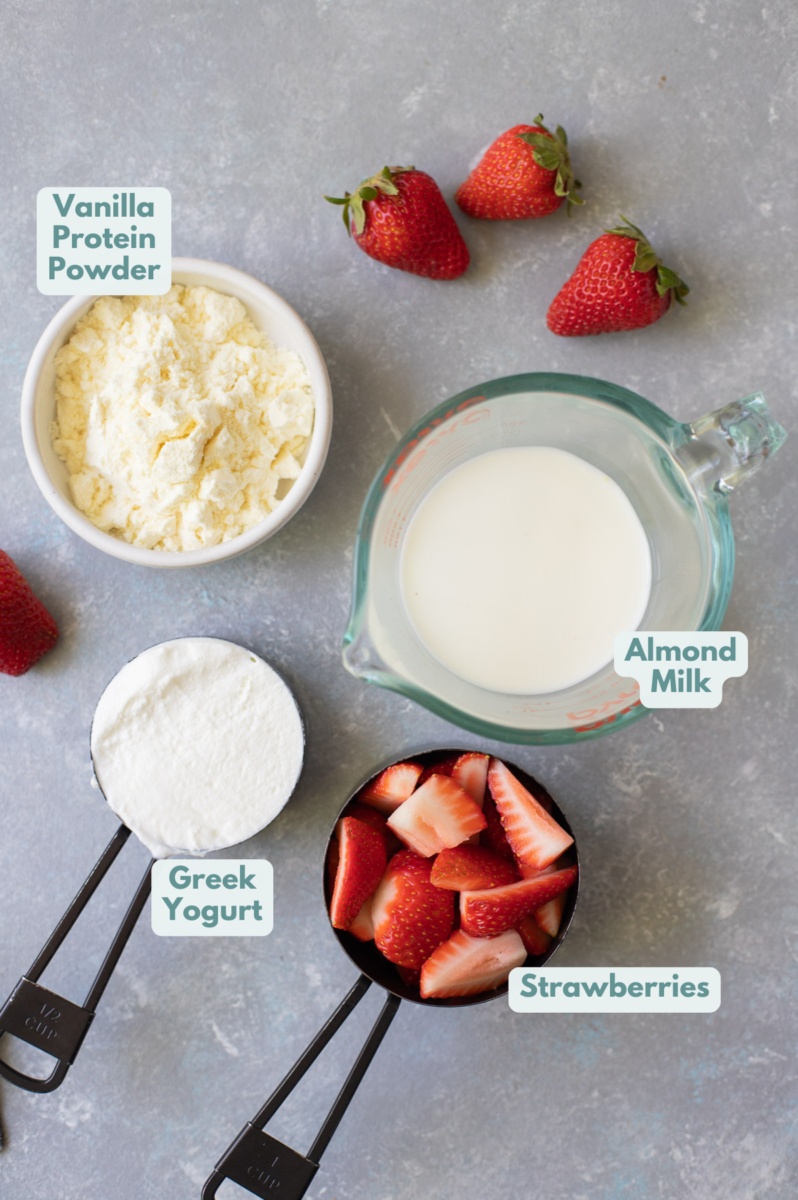 Ingredients for strawberry smoothie in white bowls and measuring cups: strawberries, greek yogurt, almond milk, vanilla protein powder