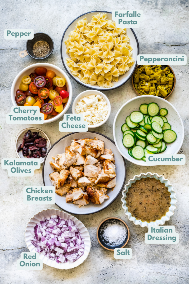 Ingredients for greek chicken pasta salad in white bowls: chicken breasts, olive oil, farfalle pasta, cherry tomatoes, cucumber, red onion, pepperoncini, kalamata olives, feta cheese, red wine vinegar, lemon juice, dijon mustard, dried oregano, salt and pepper