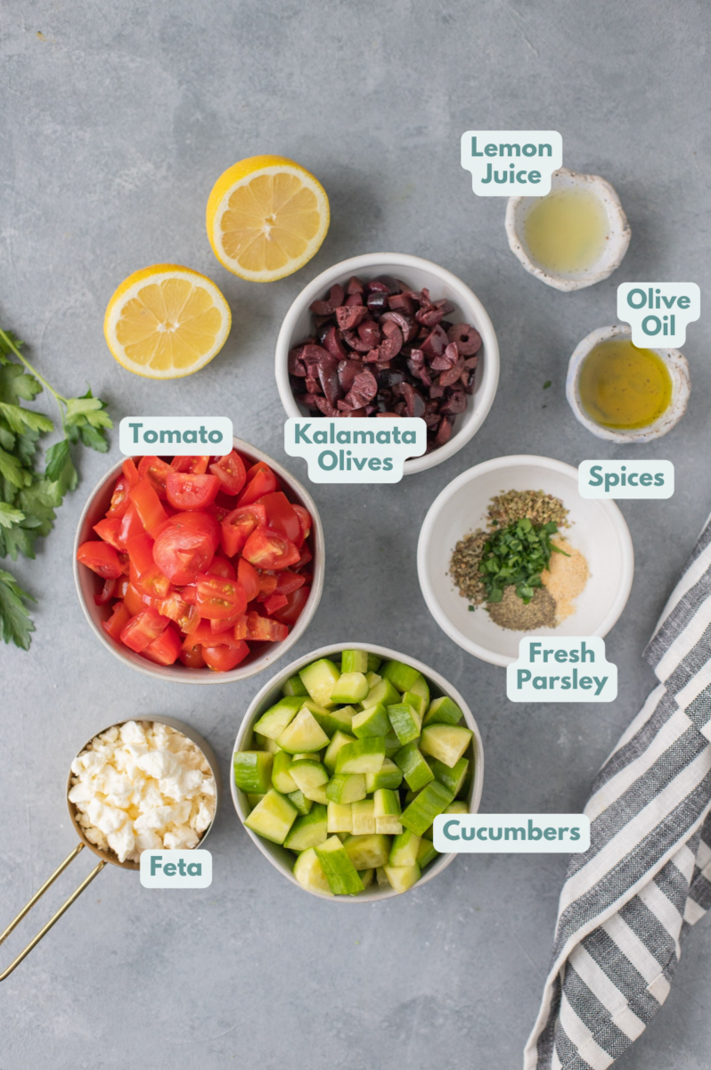 Ingredients for Greek Salad in small white bowls: chopped tomatoes, kalamata olives, cucumbers, feta cheese, lemon juice, olive oil, parsley, garlic powder, dried oregano, dried basil