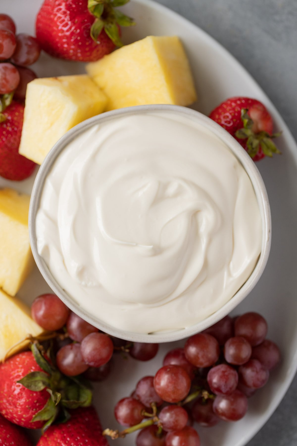 Overhead view of healthy fruit dip in a white bowl on a platter, surrounded with fresh grapes, strawberries and pineapple chunks.