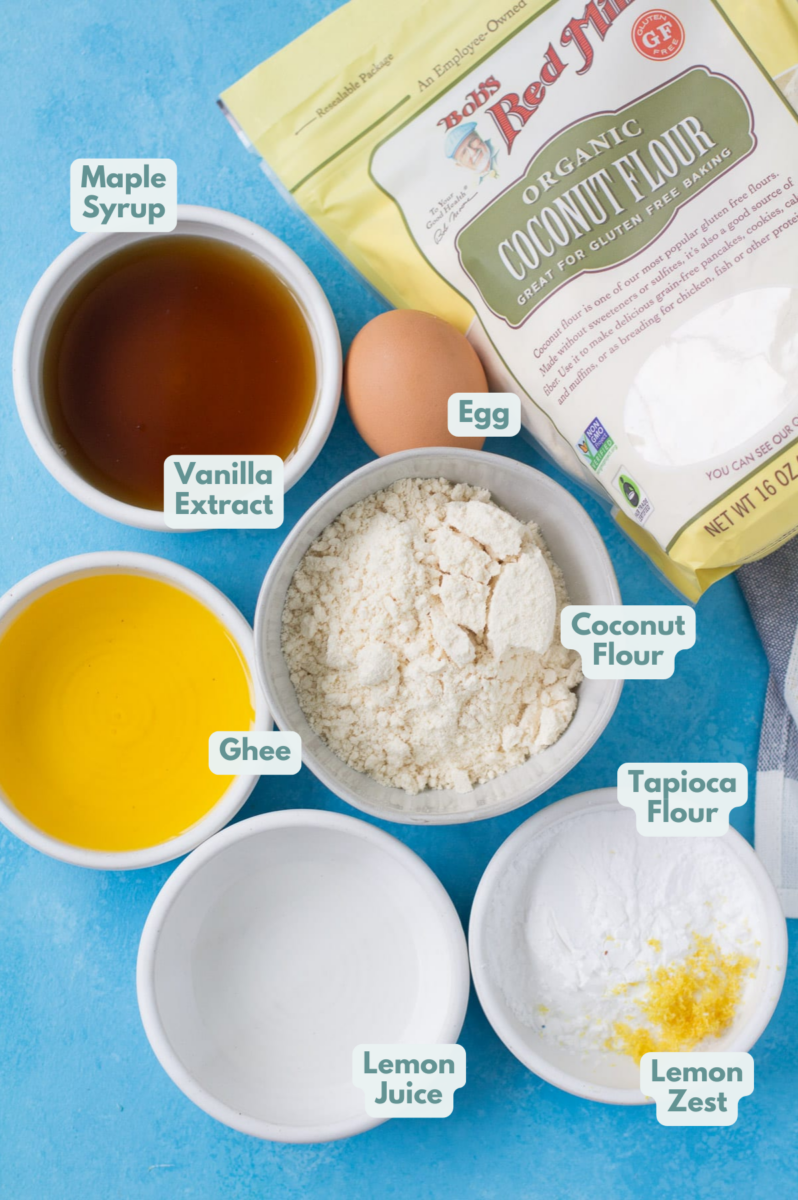 Ingredients for healthy lemon bars in small white bowls on a bright blue background - maple syrup, lemons, egg, melted ghee, coconut flour, tapioca flour.