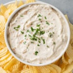 An overhead shot of a bowl of Healthy Onion Dip with potato chips on the side on a grey background