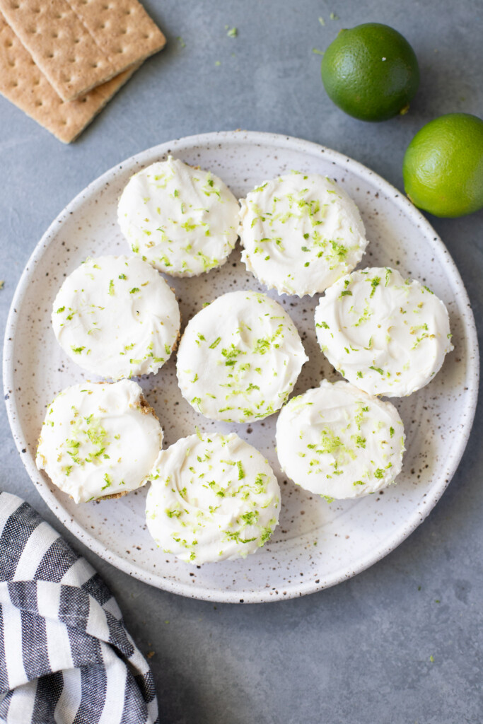 An overhead shot of a white plate of key lime cheesecakes on a grey background with limes on the side