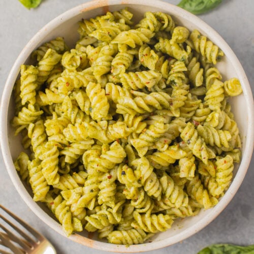 An overhead shot of a white bowl of pasta with nut free pesto and basil leaves on the side