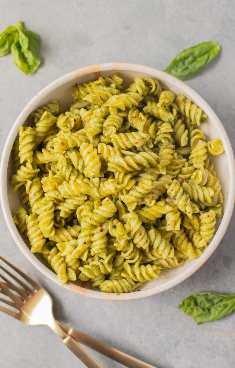 An overhead shot of a white bowl of pasta with nut free pesto and basil leaves on the side