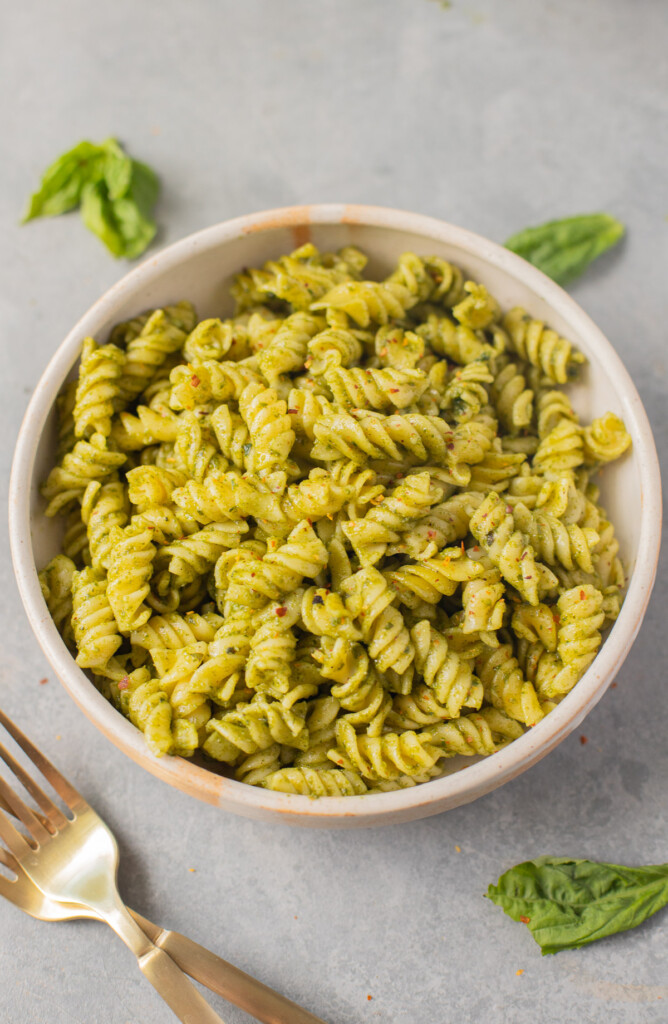 A white bowl of pasta with nut free pesto and basil leaves and a fork on the side
