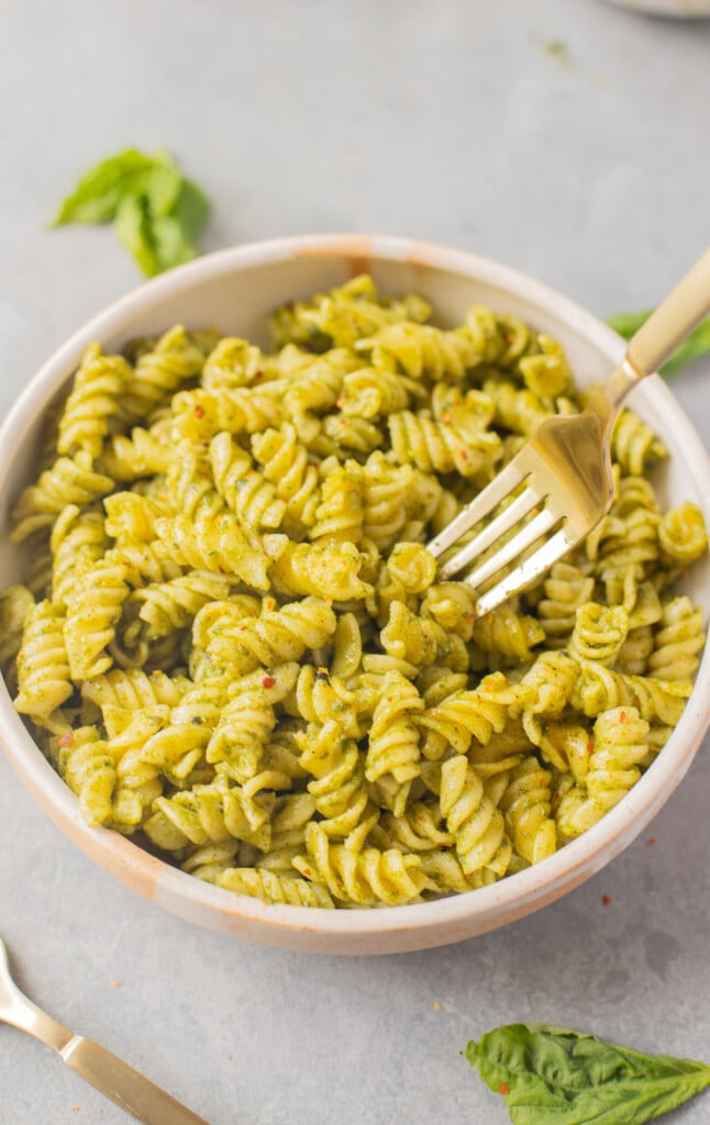 A white bowl of pasta with nut free pesto with a fork taking a bite out of it and basil leaves on the side