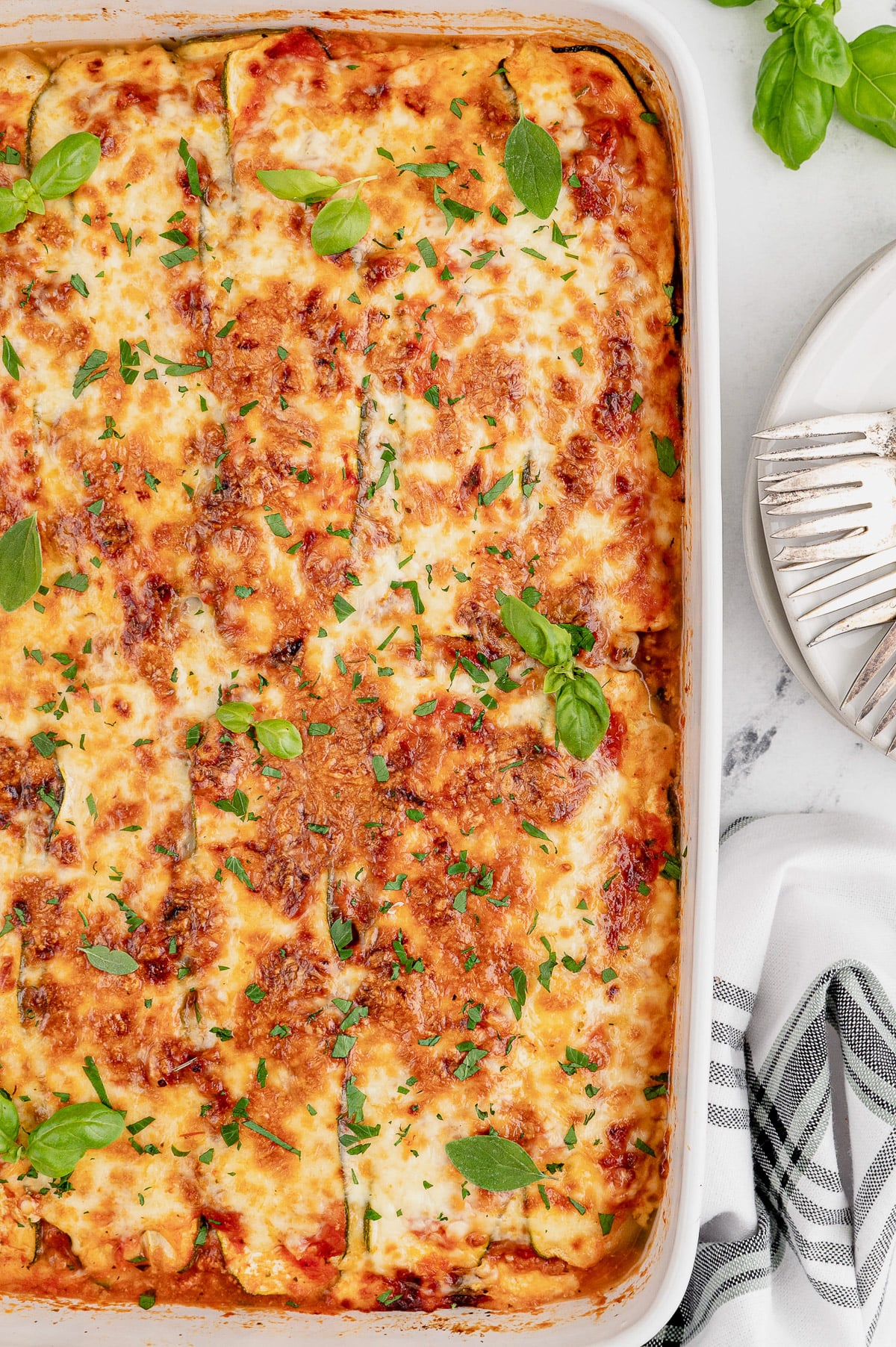 An overhead shot of a white casserole dish of zucchini lasagna with fresh basil on top on a marble counter