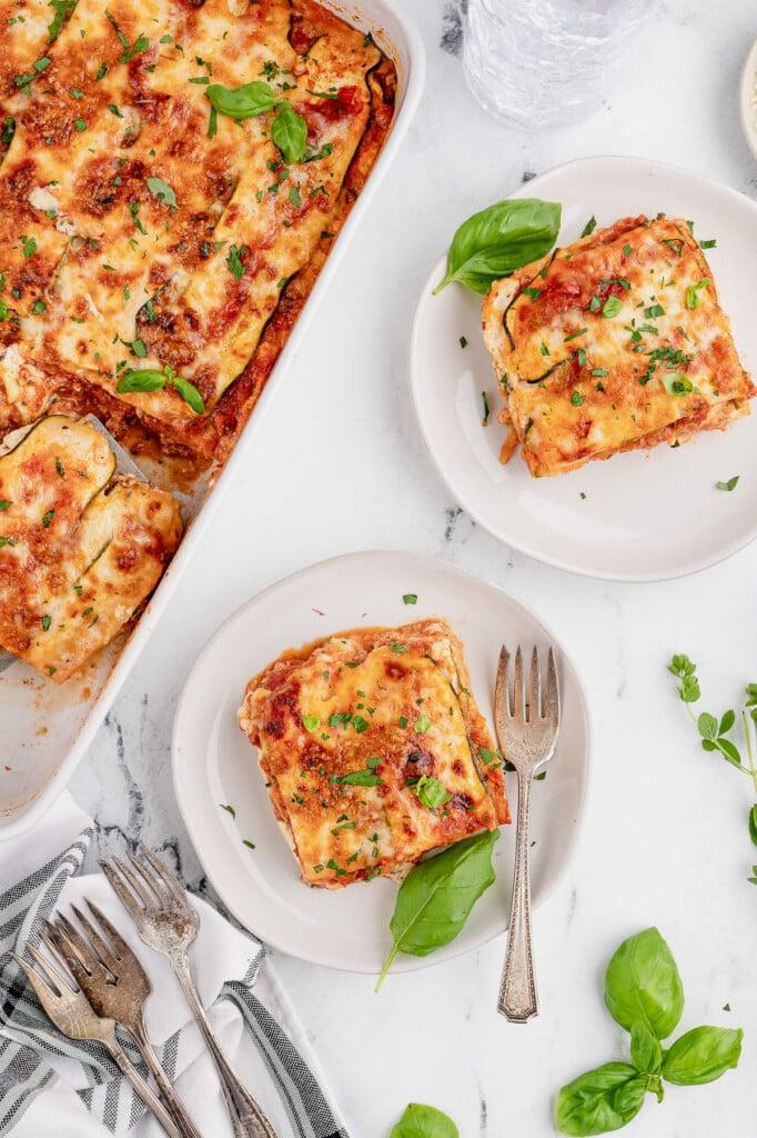 An overhead shot of two white plates with pieces of zucchini lasagna and a white baking dish on the side