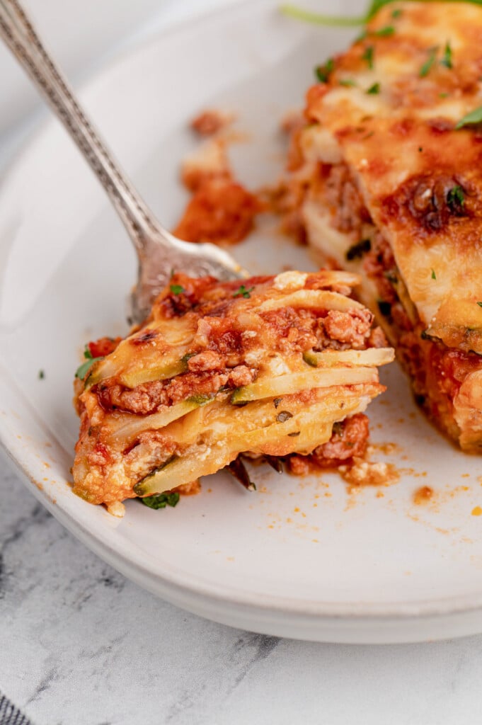 A fork with a bite of zucchini lasagna from a piece on a white plate